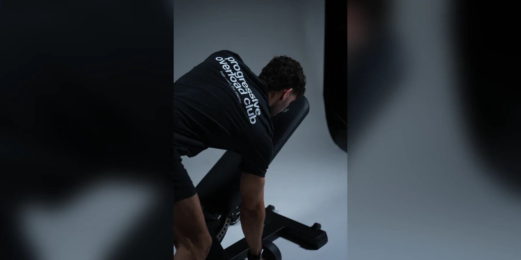 An athlete resting over a weight bench in a "progressive overload club" shirt